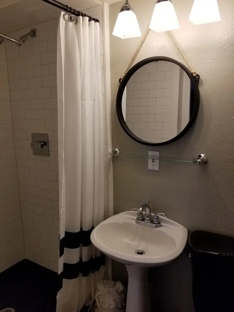 Small bathroom with white subway-tile shower, dark walls, a round mirror and a pedestal sink