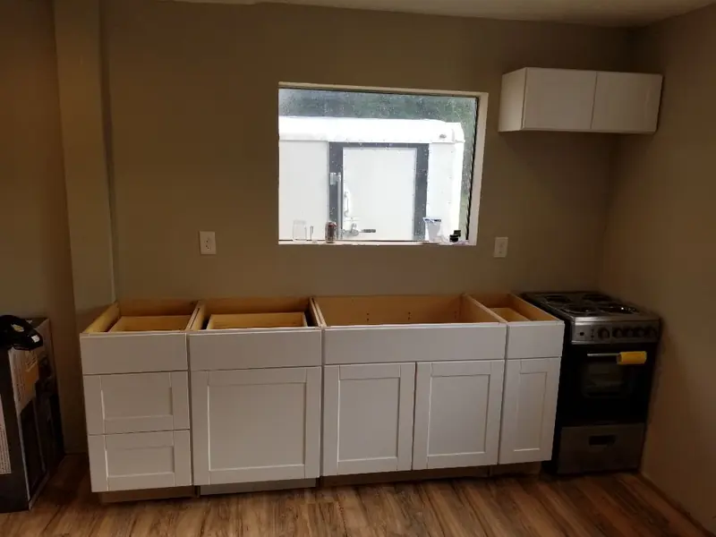 Row of new white shaker base cabinets being set beside a range, before the countertop goes on