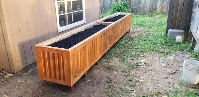 Long cedar raised planter box filled with soil along a backyard fence