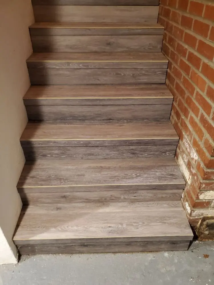 Stair treads and risers covered in grey wood-look vinyl plank beside a brick wall